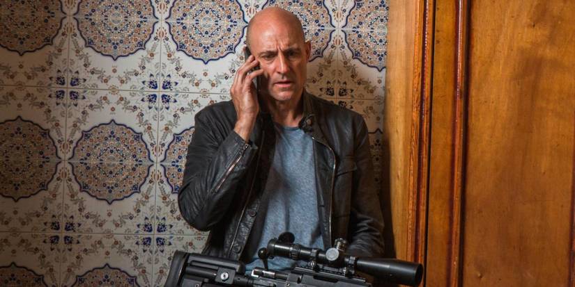 Mark Strong sitting down with a rifle in his lap while on the phone in Deep State 