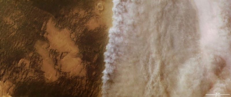 Large dust storm seen from above over reddish terrain, with a turbulent leading edge of white, puffy-looking dust.