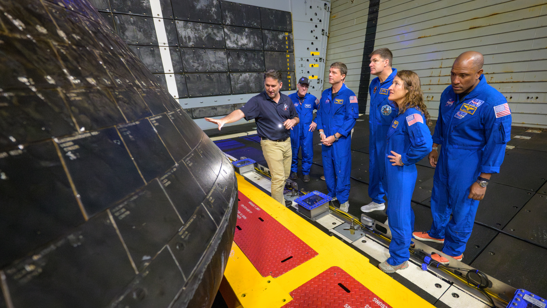 Four people wearing blue jumpsuits stand next to a person pointing to a black, round heat shield.