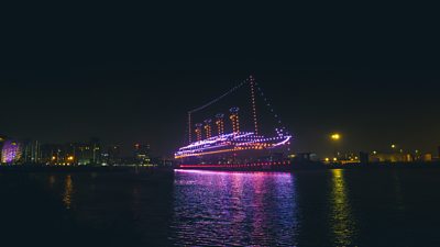 A large light display of a ship, outlined with colourful neon lights made of drones, is illuminated above the water in Belfast harbour. There is a pink reflection on the water and there are city lights in the background.