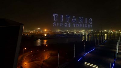 Image shows a nighttime cityscape with bright lights reflecting on the water. Above the harbour, a large formation of illuminated drones spells out the words “TITANIC SINKS TONIGHT” against the dark sky.
