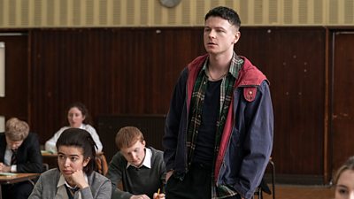 Young Ruben (Stuart Campbell) stands in an exam hall in casual clothes. Students in school uniforms sit at desks around him. 