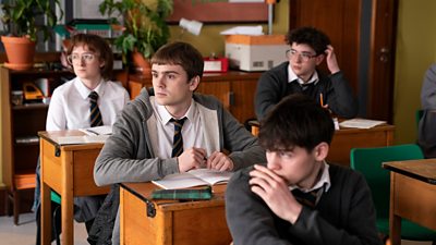 Young Niall (Mitchell Robertson) sits at a desk in school uniform. Other students sit at desks in the classroom around him. 