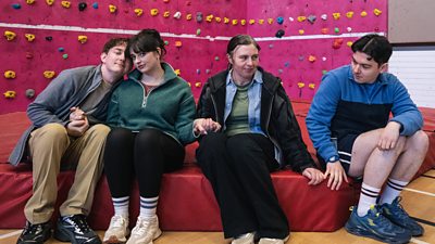 Four people sit on the edge of a safety mat below a climbing wall. Three people hold hands on the left of the image, while a fourth watches. 