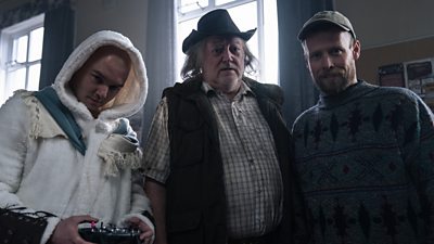 Three people stand an a large room and look down at the camera. One holds a large remote controller. 