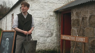 A man holds a bucket outside a period building. Signage beside the buidling reads: "Interactive exhibit." 