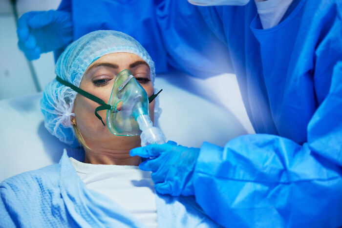 Patient under sedation wearing oxygen mask while surgical staff in protective gear administer anesthesia before surgery.