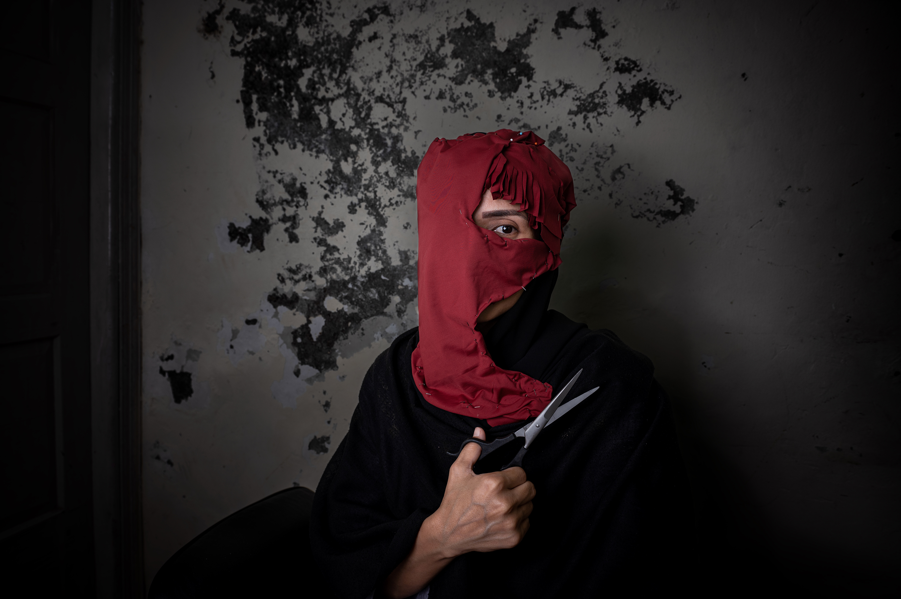 A person with their face almost entirely covered by a torn red headscarf holds a pair of scissors against their chest, gazing through the gap with a single visible eye against a dark, peeling wall.