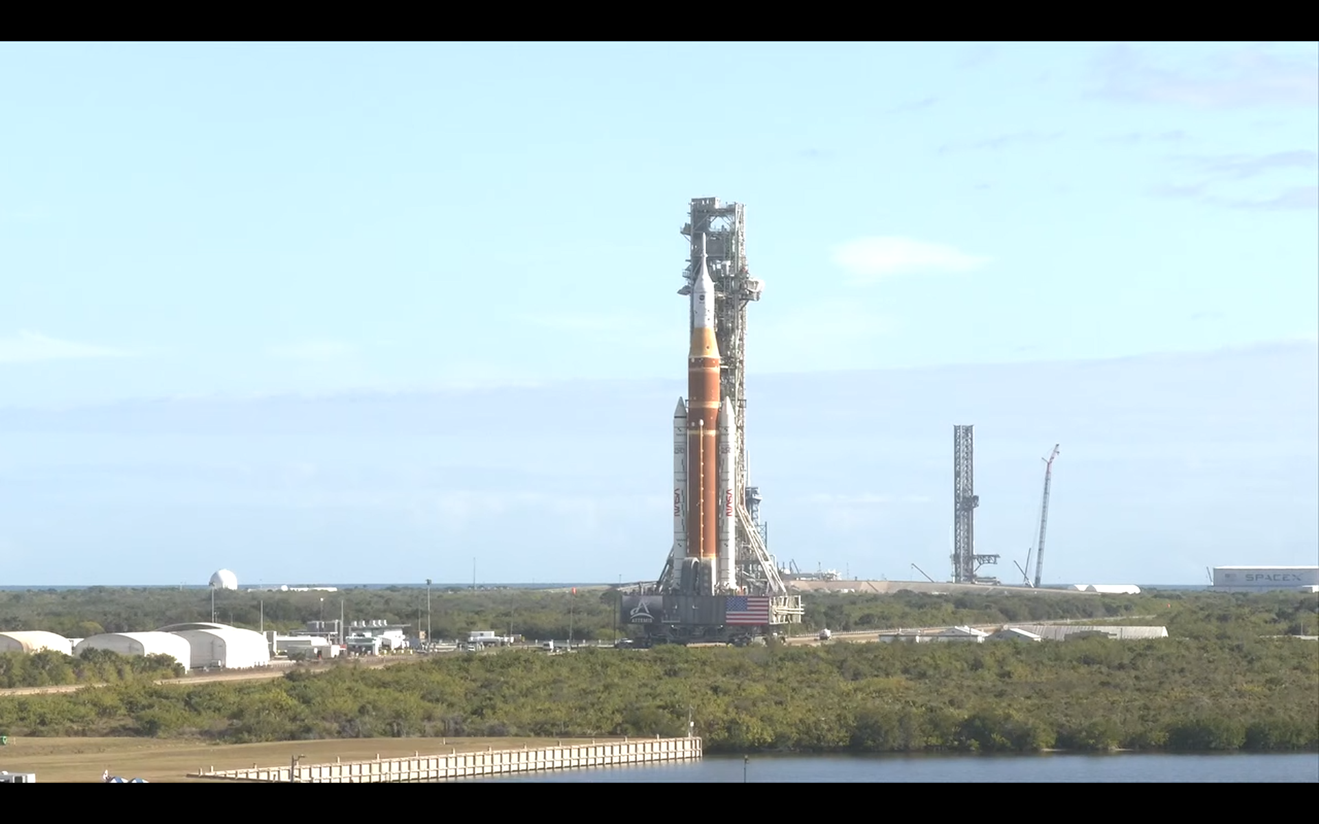A towering NASA Artemis 2 moon rocket with twin white side boosters being hauled toward a seaside launch pad.