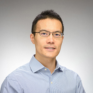 A smiling East Asian man with short dark hair and rectangular glasses wears a blue and white striped collared shirt against a grey background.