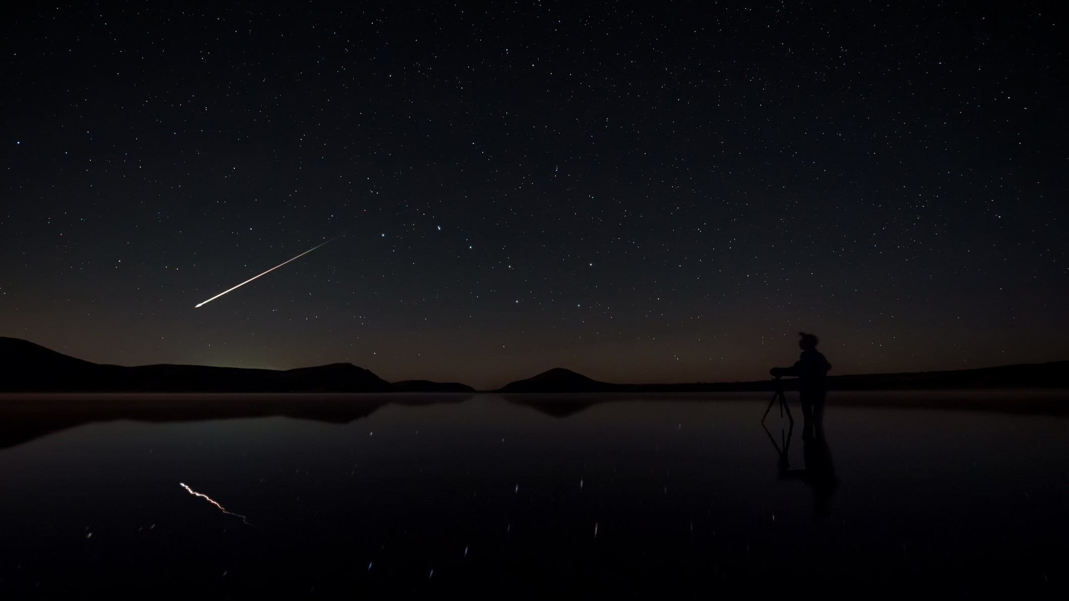 a bright shooting star streaks across the sky and is reflected in the water below.