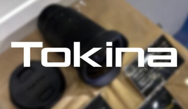 A blurred photo of a camera lens and lens cap on a table, with the word "Tokina" in bold white letters prominently overlaid in the center.
