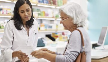 A pharmacist in a white coat reviews a prescription while consulting with an elderly woman at a pharmacy counter.