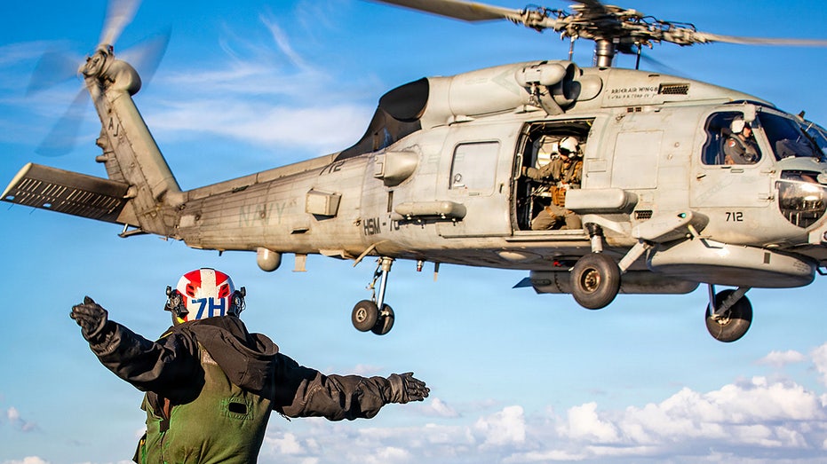U.S. Navy sailor signals helicopter launch on aircraft carrier flight deck.