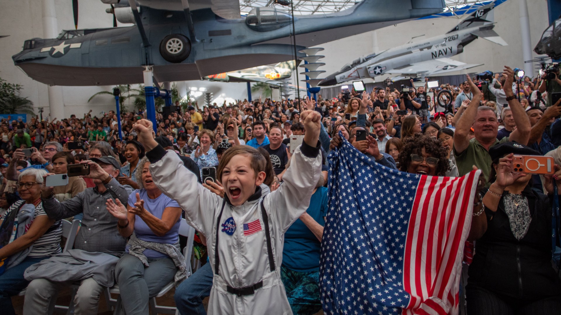 A young boy wears an astronaut costume next to a woman waving a flag as they watch the return of the Artemis 2 crew members to Earth at the San Diego Air and Space Museum on April 10, 2026.