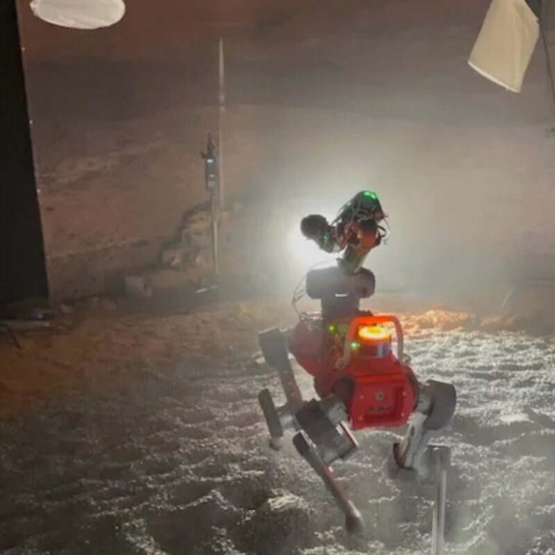 Dog-like robot with a red body and 4 legs on gravel indoors. It has a bright headlight.
