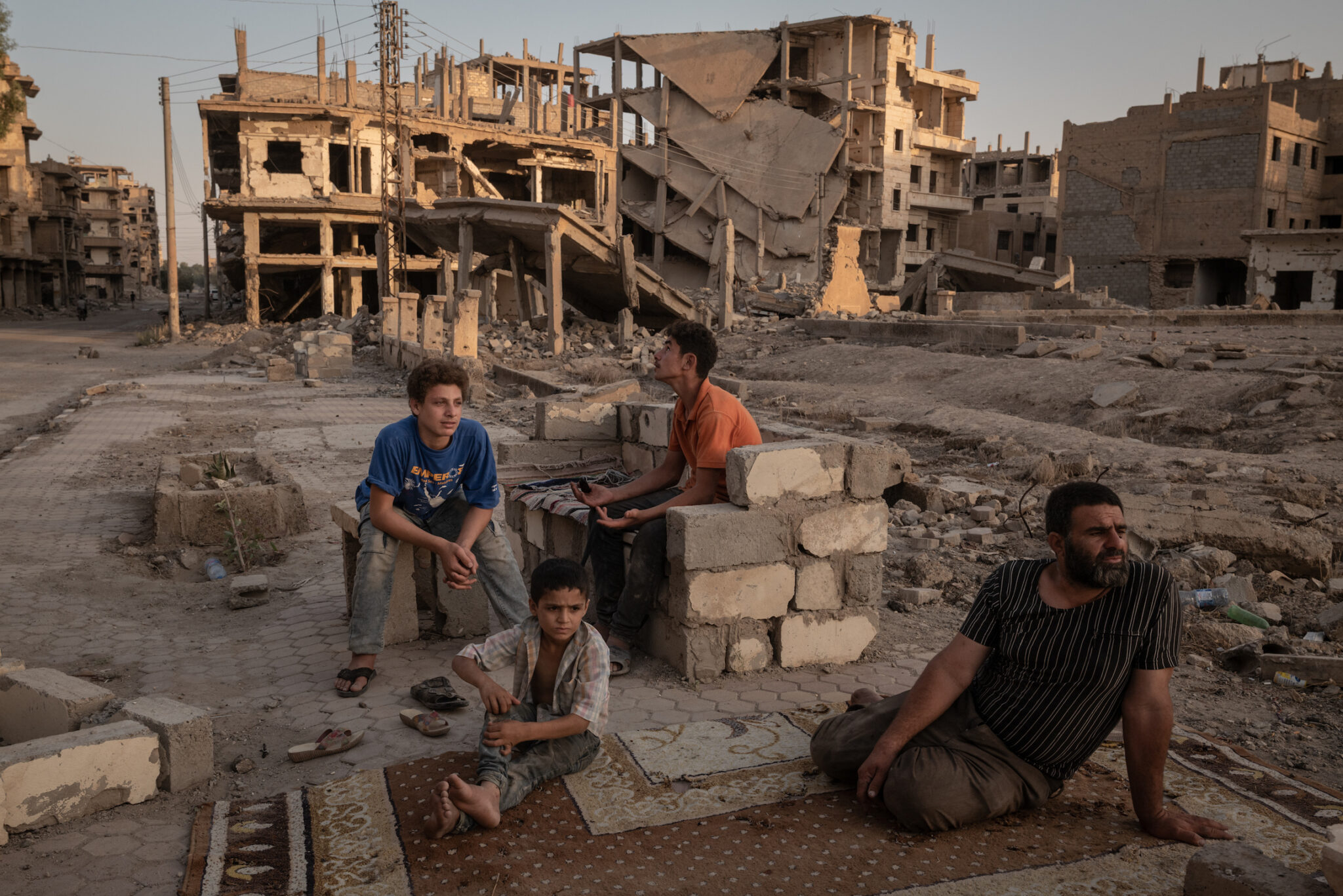 Nicole Tung, “A Syrian City Rebuilds, Still Divided,” for the New York Times. Abdelatif Daham Al Hummada (r.) sits with his sons and nephew on the street outside their heavily damaged home, where the family often sleeps. Deir al-Zour, Syria, August 20, 2025.