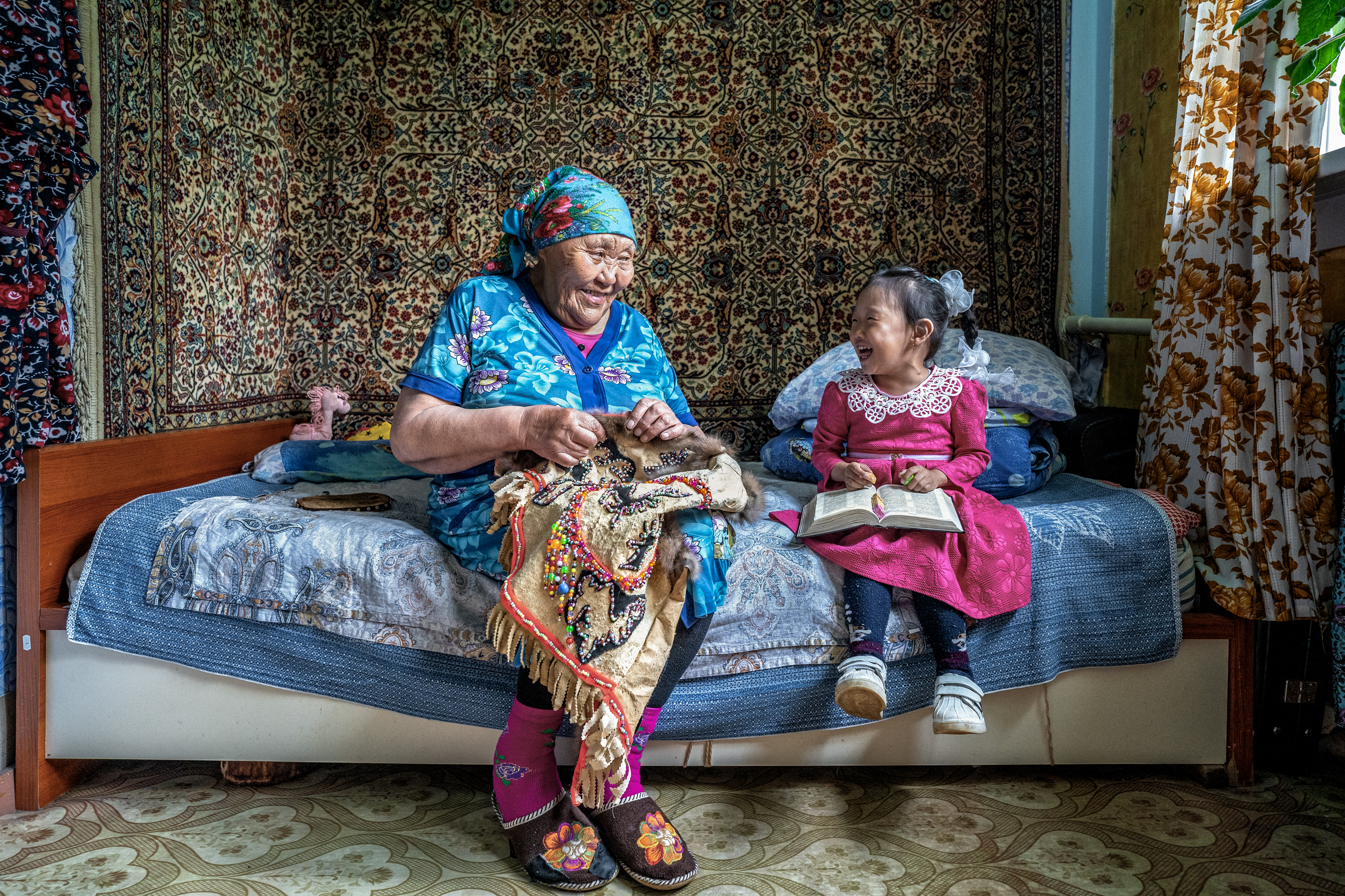 An elderly woman sits on a bed sewing a richly decorated textile while smiling at a young girl beside her, who holds an open book, both sharing a warm moment in a cosy, patterned room.