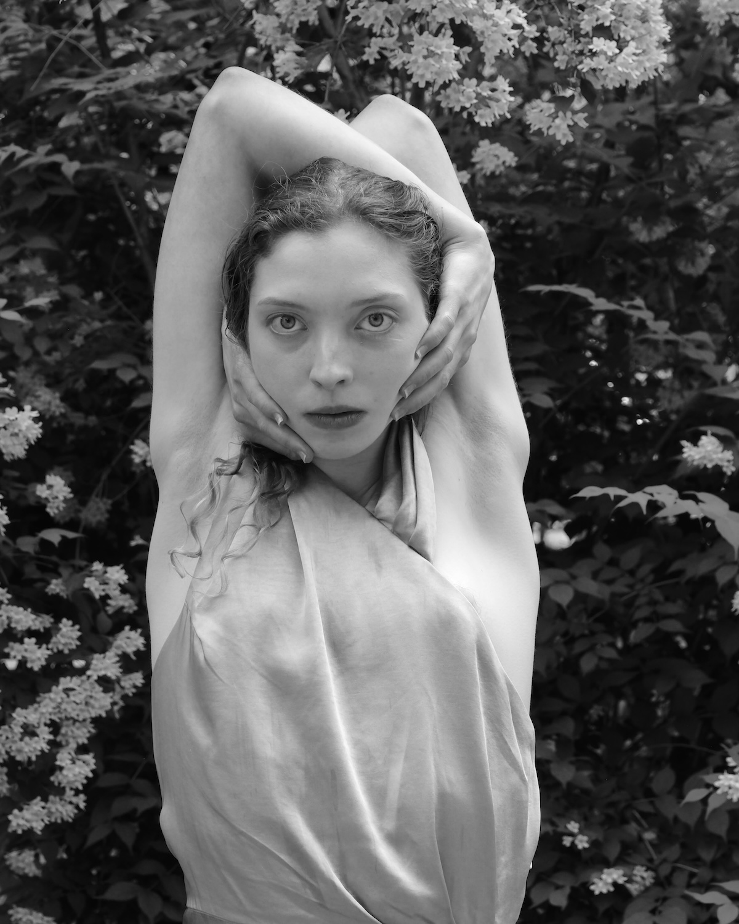 A young woman with her arms raised above her head gazes directly at the camera in a black-and-white portrait taken among blooming garden shrubs.