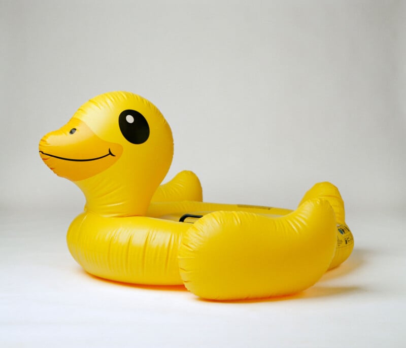 A bright yellow inflatable pool float shaped like a duck, with a smiling face, set against a plain white background.
