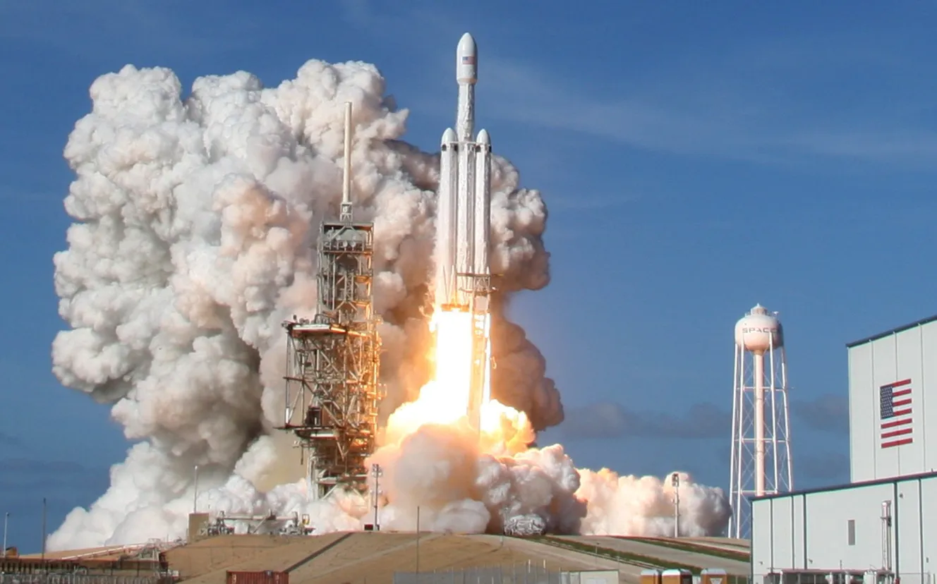 SpaceX Falcon Heavy rocket lifting off from Launch Pad 39-A at Kennedy Space Center, with bright orange flames and white smoke billowing around it.
