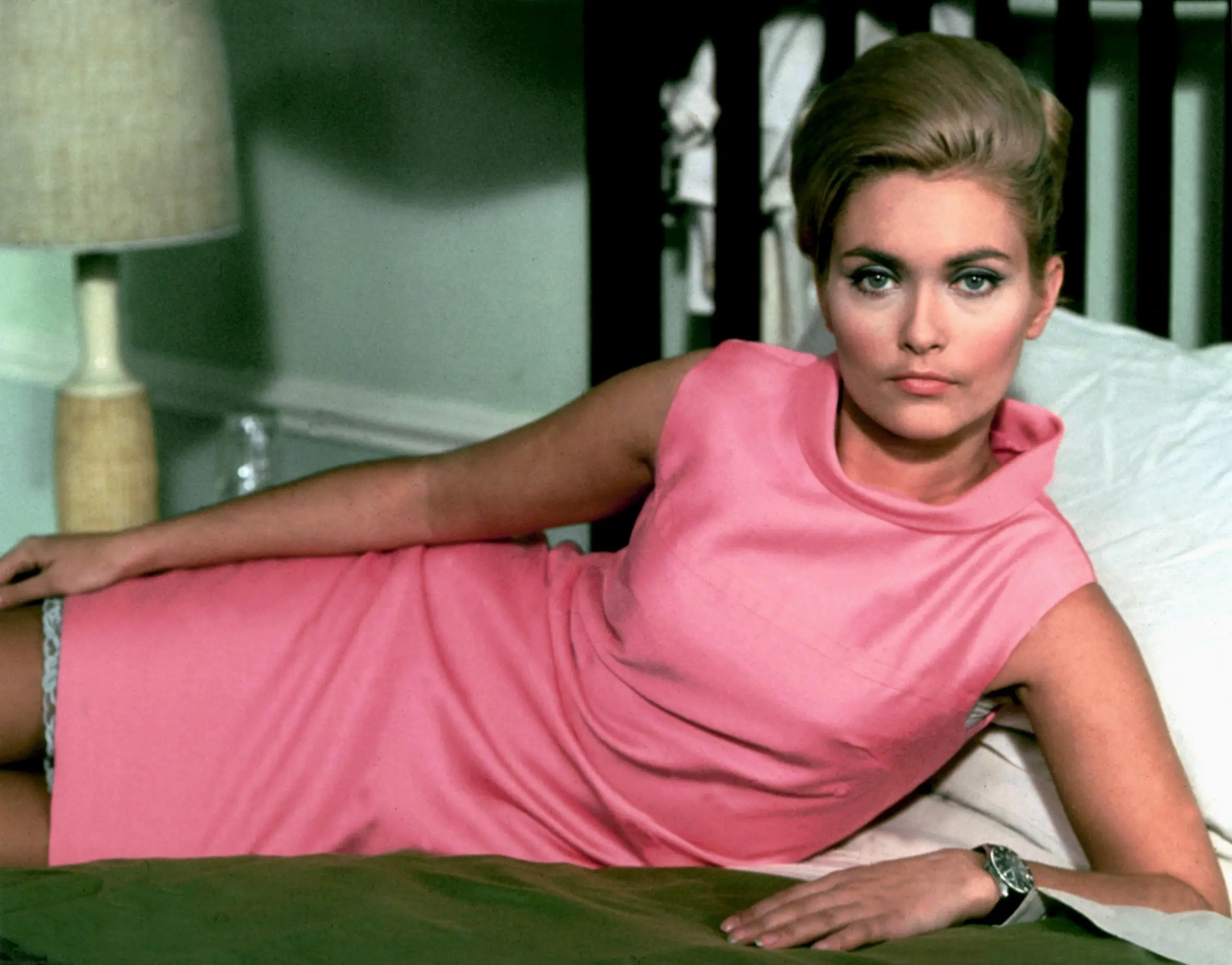 Alexandra Bastedo as Sharron Macready in "The Champions" in a pink dress, lying on a bed with a lamp in the background.