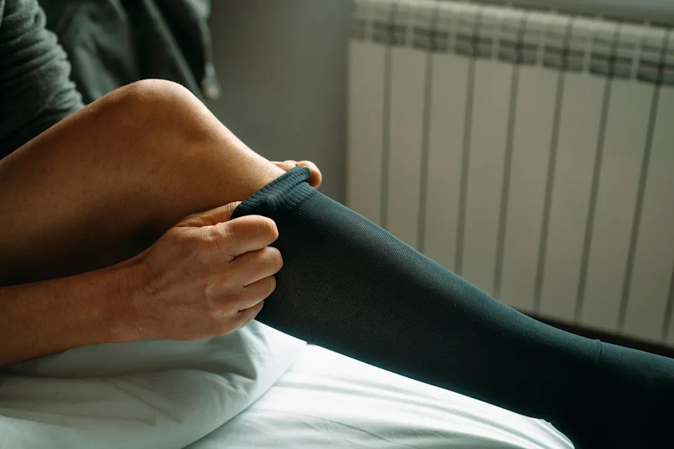 Person putting on a knee-high black sock, sitting on a bed. Close-up of hands pulling the sock up over the leg