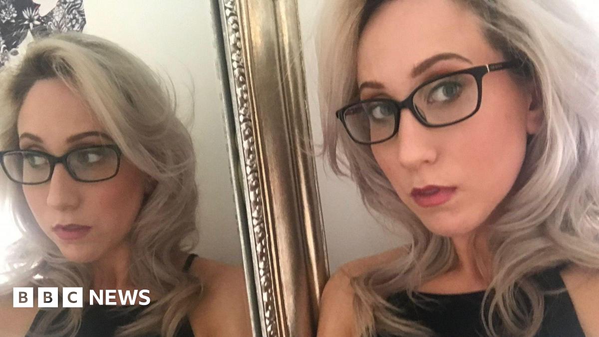 A woman with blonde hair and dark rimmed glasses stands next to a gold rimmed mirror which shows her full reflection
