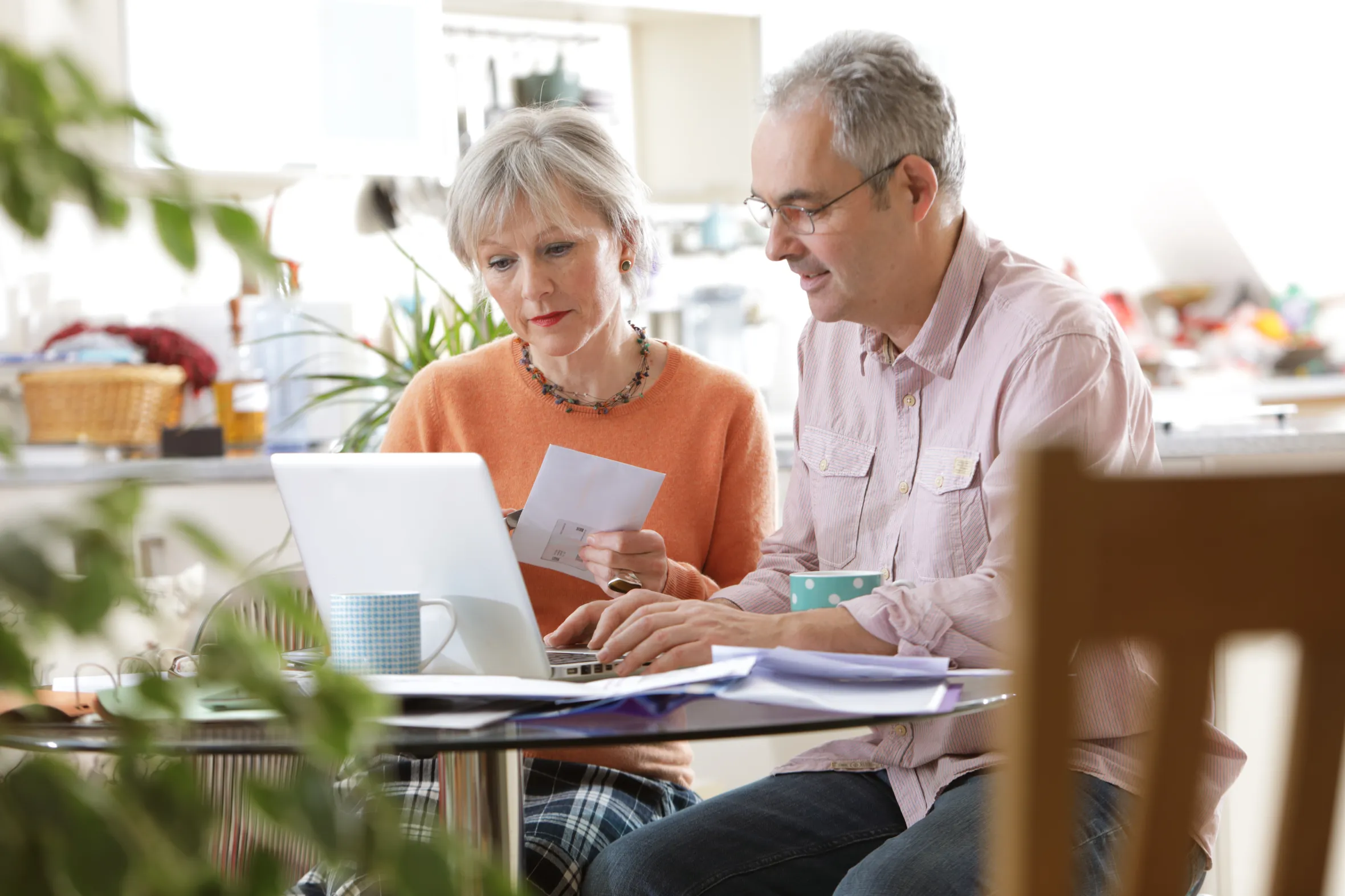 An older couple sits at a glass table, reviewing financial bills and using a laptop for home finances.