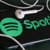 This photo illustration shows a smartphone with Spotify's green logo on its screen. White earphones with white wires are on top of the phone.