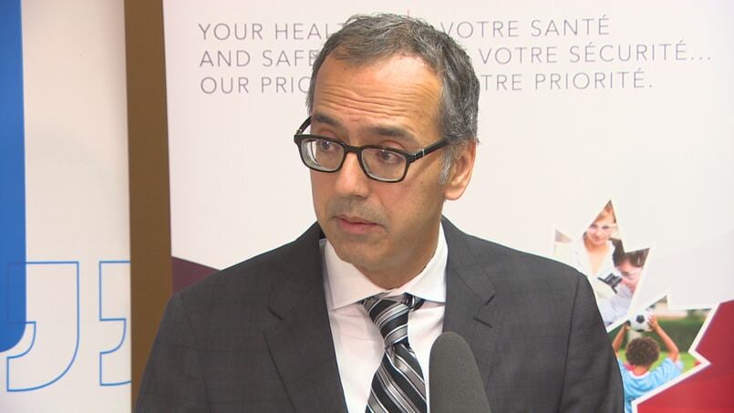 A man with short black hair and eyeglasses, wears a grey suit and striped tie