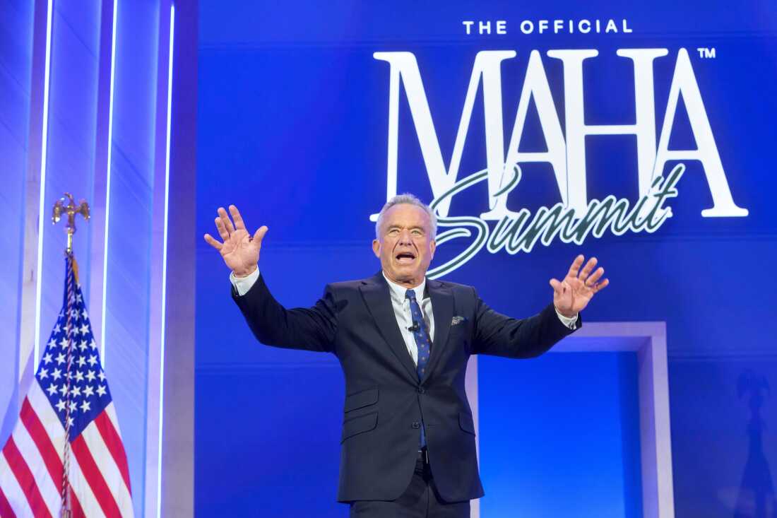 Health Secretary Robert F. Kennedy Jr. is shown with his arms raised enthusiastically while standing on a stage. The words "The official MAHA Summit" are in large print on a blue backdrop behind him.