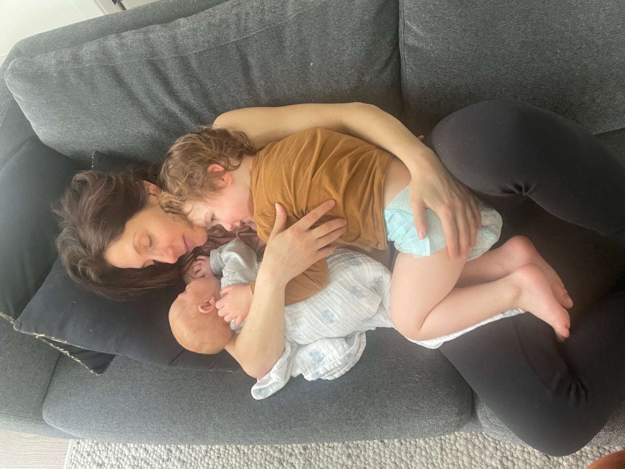 A mother holds a baby and a toddler on a couch.