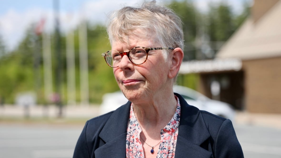 A white woman with short grey hair and glasses stands outside a municipal building with flag poles in the background. She wears a flowered shirt under a navy blazer.