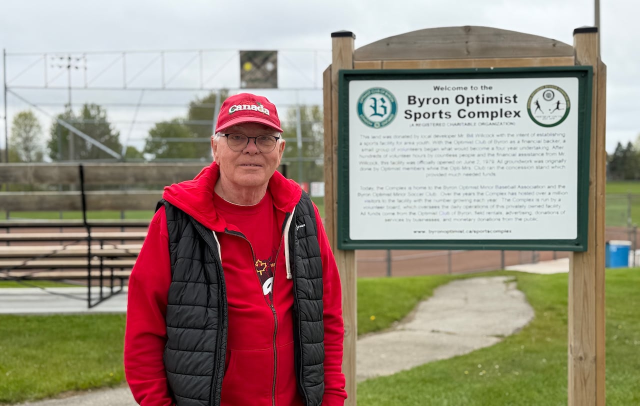 A man wearing Canada Day-themed clothing stands in front of a ballpark and sign that says "Byron Optimist Sports Complex"