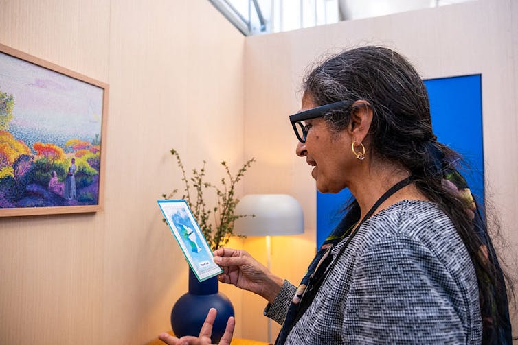 A middle-aged woman wearing glasses smiles and gazes at a large, blue tarot card in her right hand.