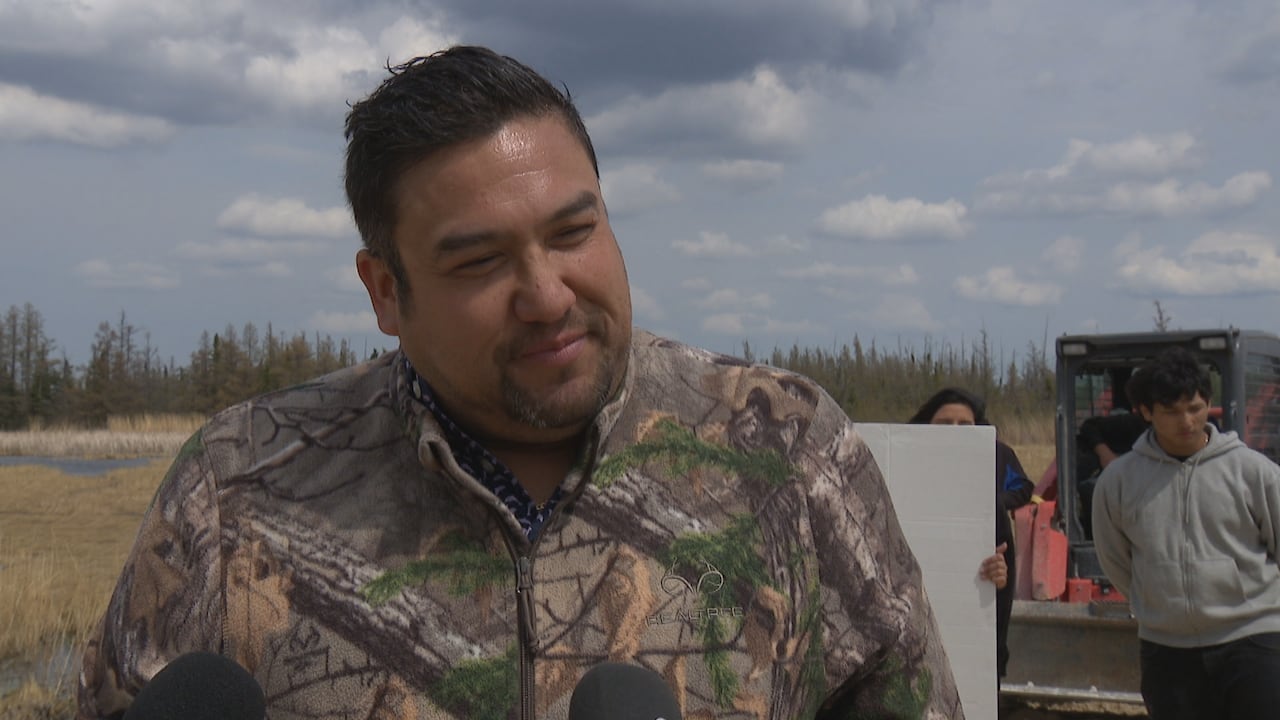A man with dark hair wears a camouflage shirt in an open marshy area, with a treeline in the background