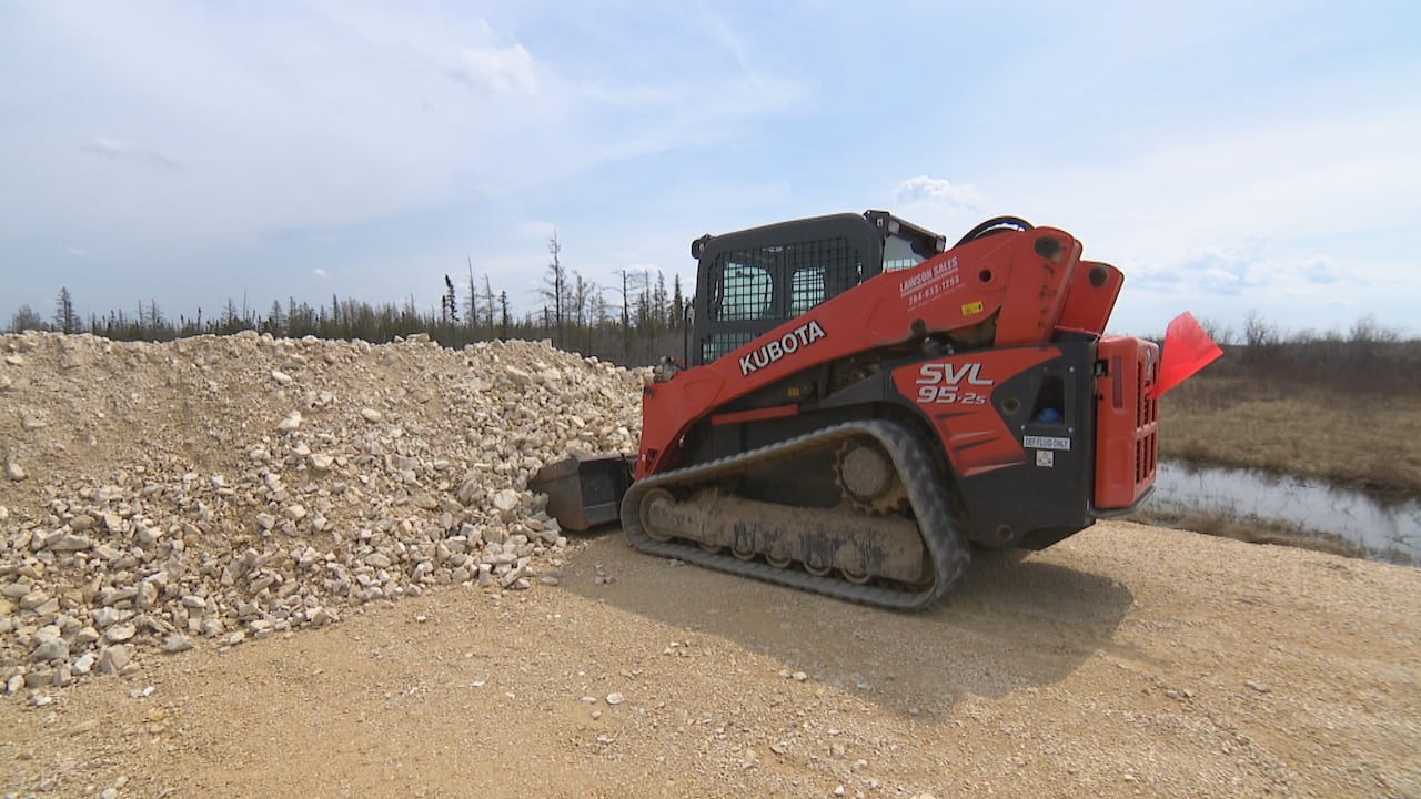 An orange track loader machine digs its bucket into a line of gravel along a road in an open marshy area, with a treeline in the background