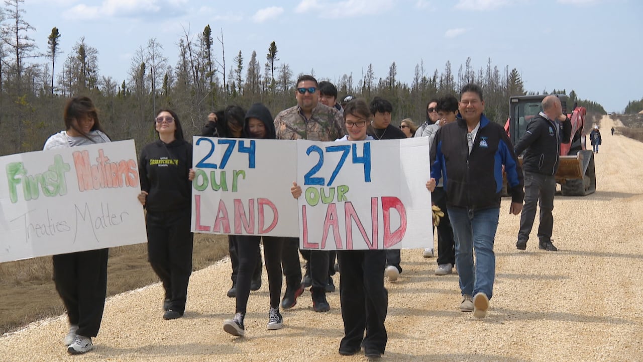 A group of people walk down a gravel road holding signs that say "274 Our Land" and "First Nations Treaties Matter"