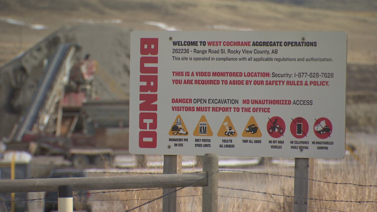 A sign on wooden posts says BURNCO and Welcome to West Cochrane Aggregate Operations, with a gravel pile in the background.