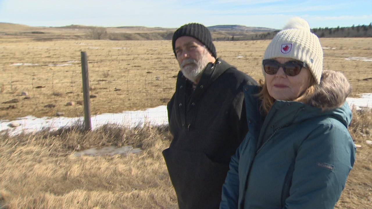 A man with a white beard wearing a black knit cap stands in a field waring a dark coat. Beside him is a red haired woman in a white knit cap wearing sunglasses.