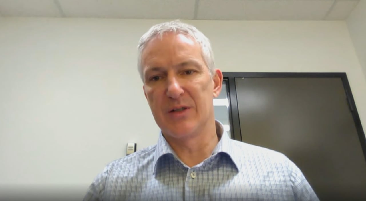 A white haired man with a collar blue striped shirt looks at the camera.