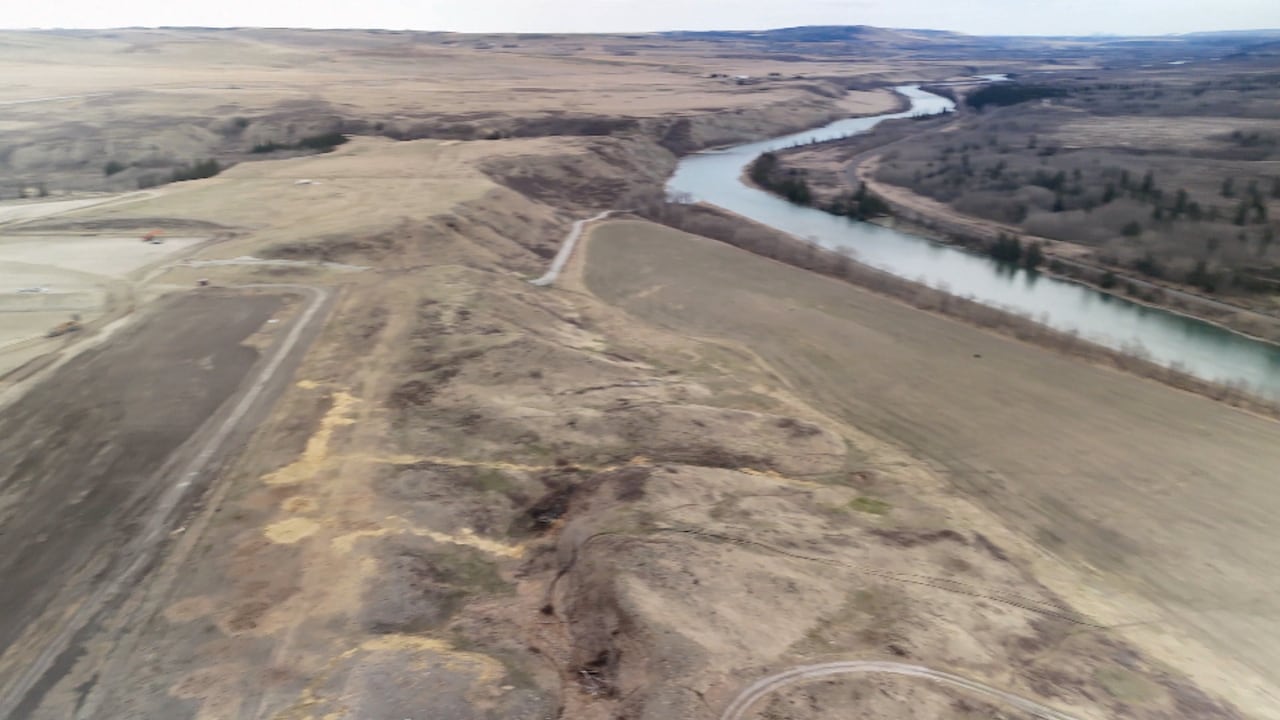 On the left is the edge of a gravel pit, on the right is a steep river bank leading down to a river in a grassland landscape with a cloudy sky.