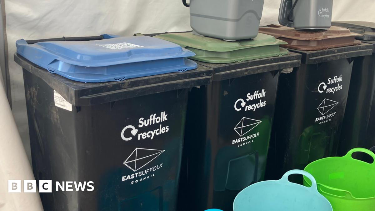 Four black bins with different coloured lids are lined up inside a tent during an event. Small grey food caddie bins sit on top of the bins.