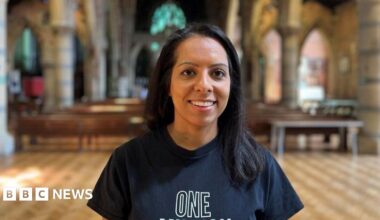 Amandeep is wearing a dark blue t-shirt, smiling at the front of a church