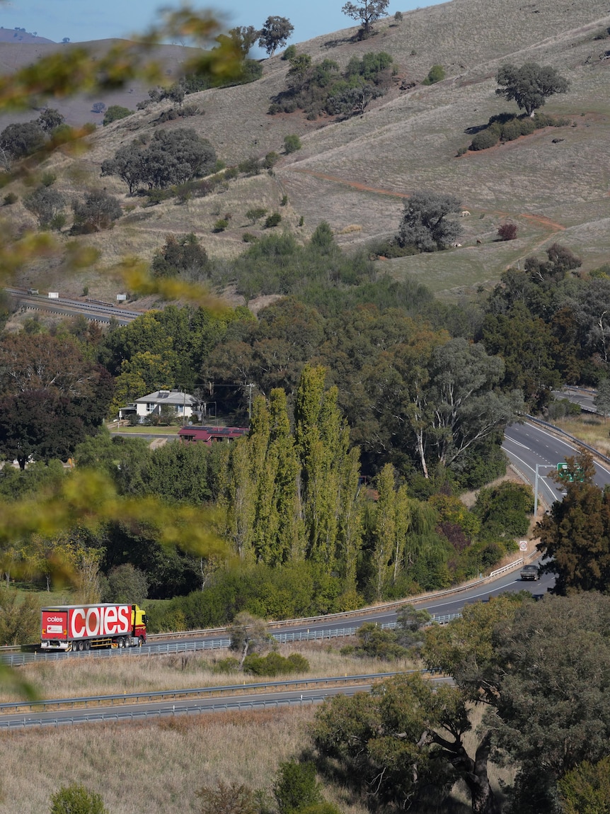 A red Coles truck drives through a winding road that curves up a grassy, green mountain.