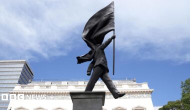 A grey statue of a man wearing a suit, walking forward off a plinth, carrying a flag that covers his face
