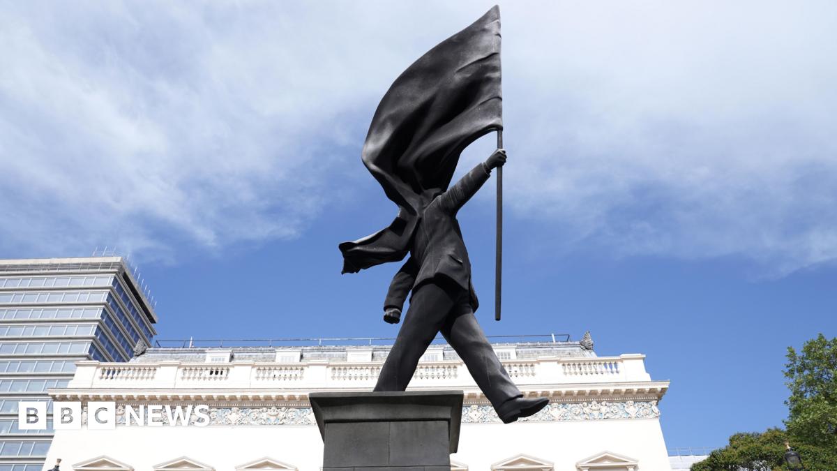 A grey statue of a man wearing a suit, walking forward off a plinth, carrying a flag that covers his face