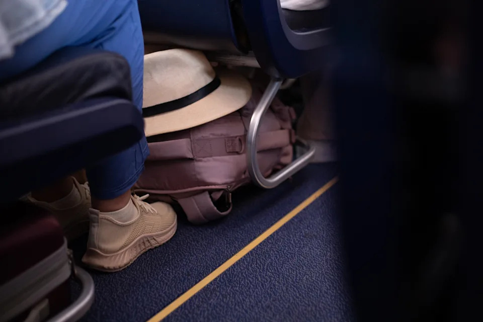 Hat and backpack under airplane seat with passenger's legs and shoes visible