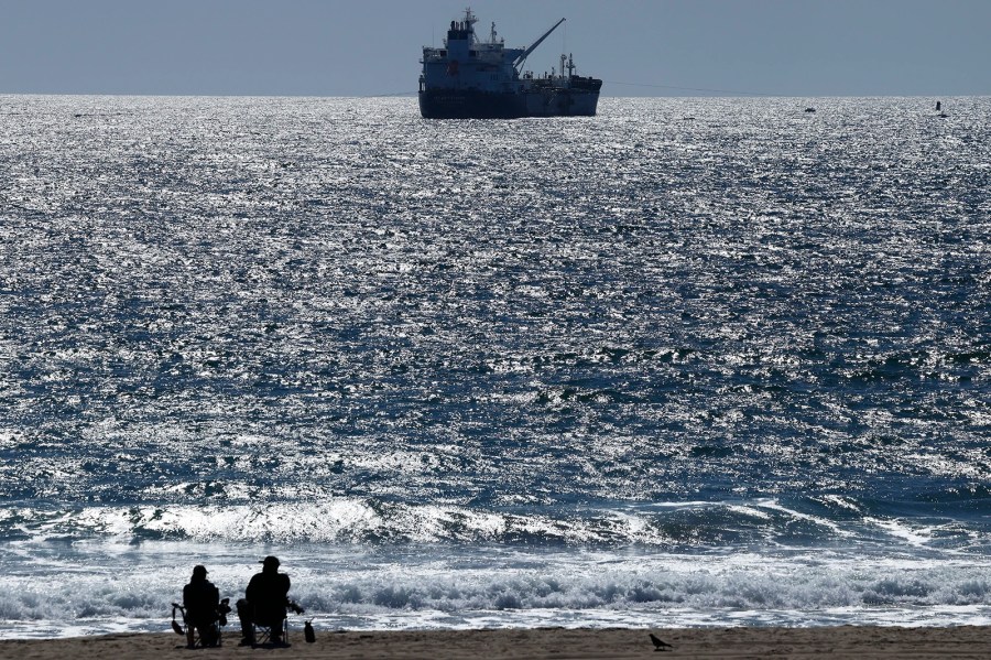 An oil tanker sits in the water near the horizon line. Two people sit on chairs on the beach.
